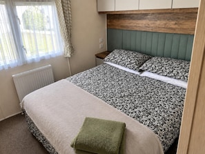 A bedroom with a bed, a nightstand, a window with curtains, and a radiator. - Grange Park 2-bed Caravan, Sleeps 6 With Parking (Mablethorpe)