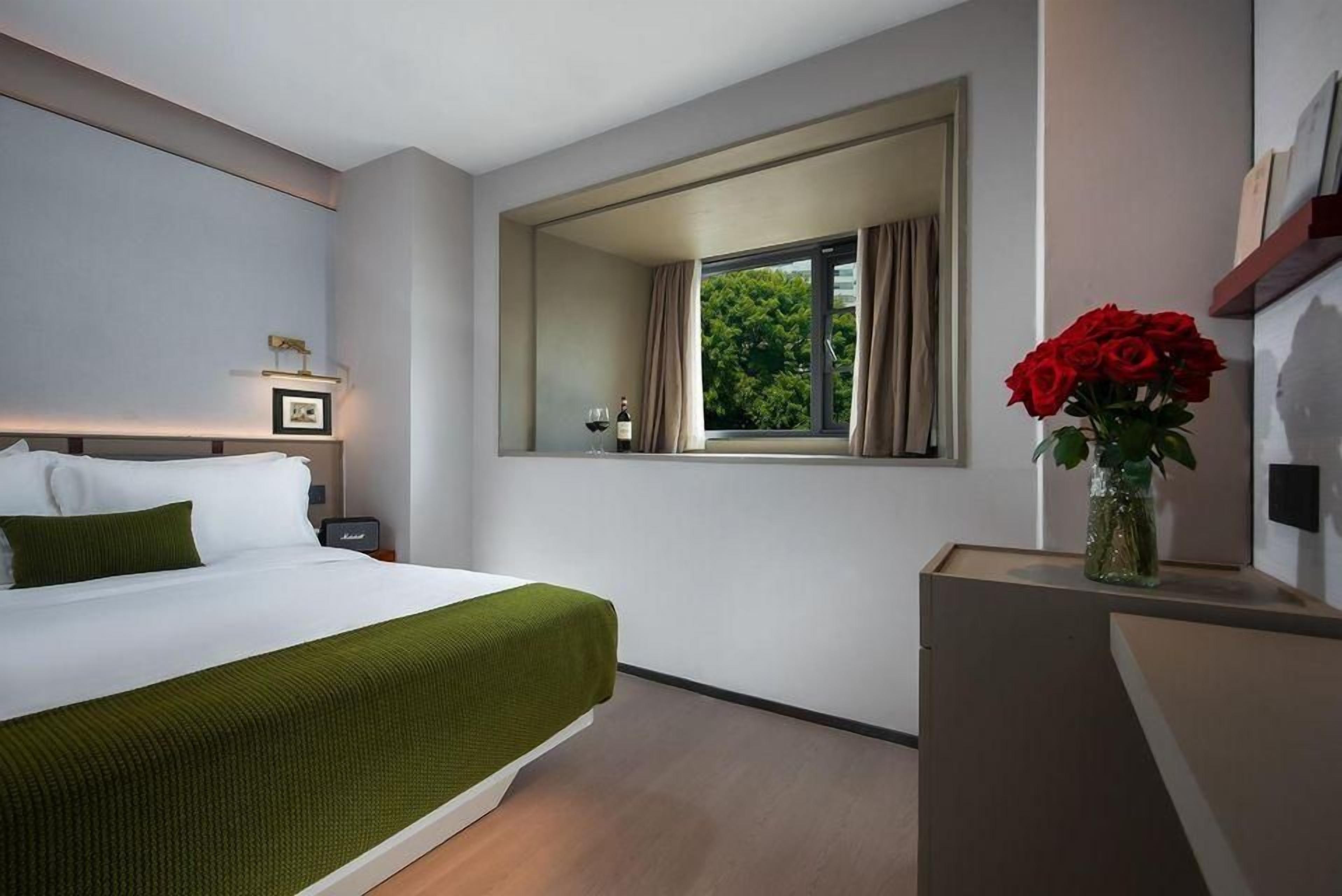 A hotel room with a bed, a nightstand, a window with curtains, and a vase with red flowers.