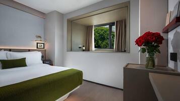 A hotel room with a bed, a nightstand, a window with curtains, and a vase with red flowers.