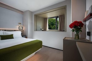 Una habitación de hotel con cama, mesita de noche, ventana con cortinas y un florero con flores rojas.