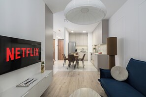 Ein modernes Wohnzimmer mit einem Flachbildfernseher, der das Netflix-Logo anzeigt, einem Essbereich mit einem Holztisch und Stühlen sowie einer Küche mit weißen Schränken und einer großen Pendelleuchte.