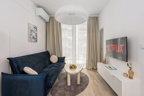 Een moderne woonkamer met een donkerblauwe bank, een klein rond tafeltje met fruit, een witte televisie waarop 'NETFLIX' staat, en een groot raam met transparante gordijnen.