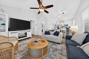 A modern living room with a flat-screen TV, a ceiling fan, a fireplace, and a dining area with a kitchen in the background. - Par For Pawleys, Golf, Pool & Near the Beach (Pawleys Island)