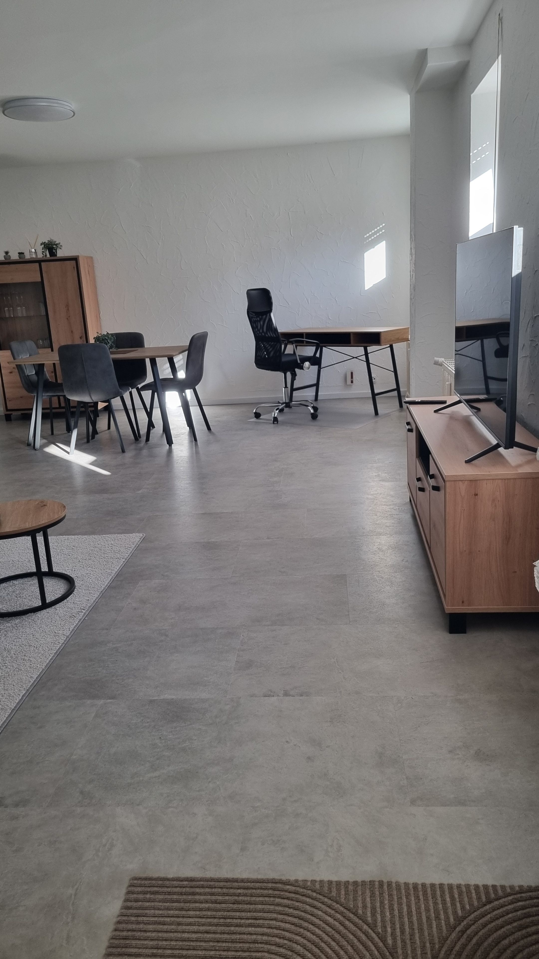 Ein moderner Arbeitsraum mit einem langen Tisch, mehreren Stühlen, einem Fernseher auf einer Holzkommode und einem gemusterten Teppich auf dem Boden.