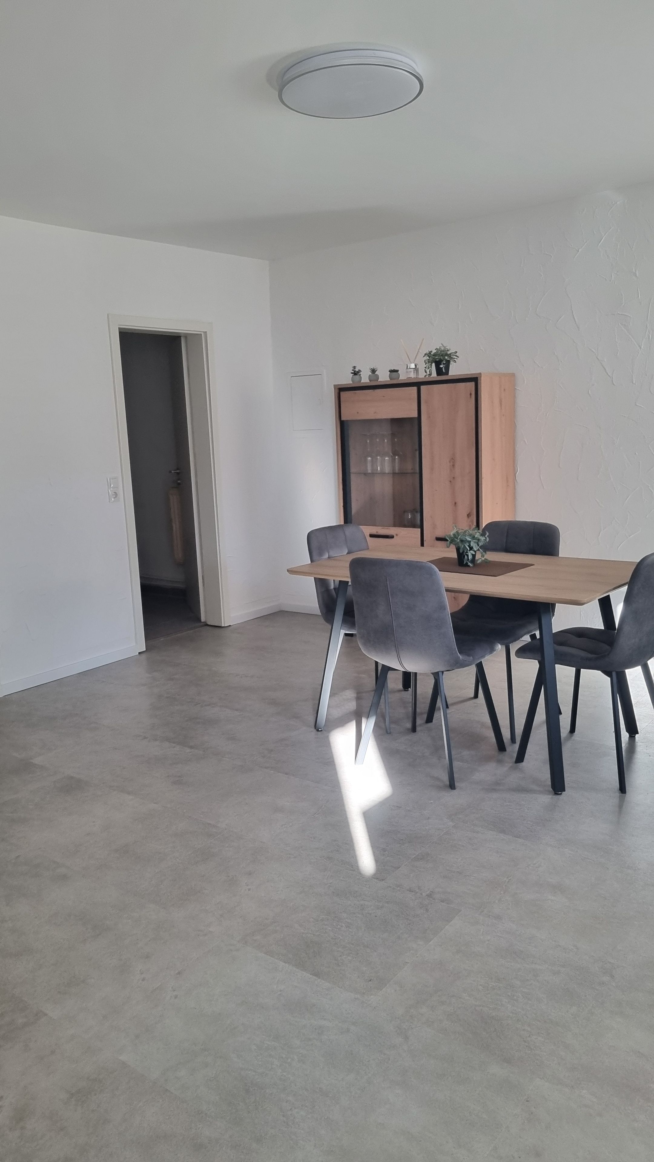 Ein Essbereich mit Holztisch und grau gepolsterten Stühlen, einem Holzschrank mit Glastüren und einer Deckenleuchte.