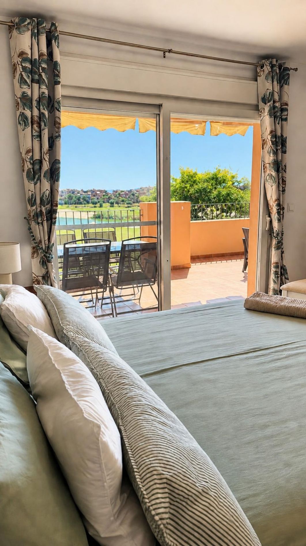 Ein Schlafzimmer mit einem Bett, einem Fenster und Blick auf einen Balkon mit Gartenmöbeln.