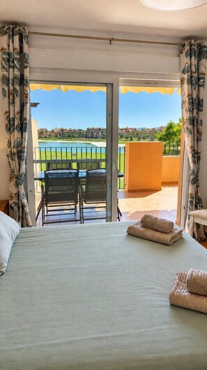 A hotel room with a bed, a view of a golf course, and a balcony with a table and chairs. - Mar Menor Golf Resort 2-Bedroom Apartment | Golf & Lake Views (MC)
