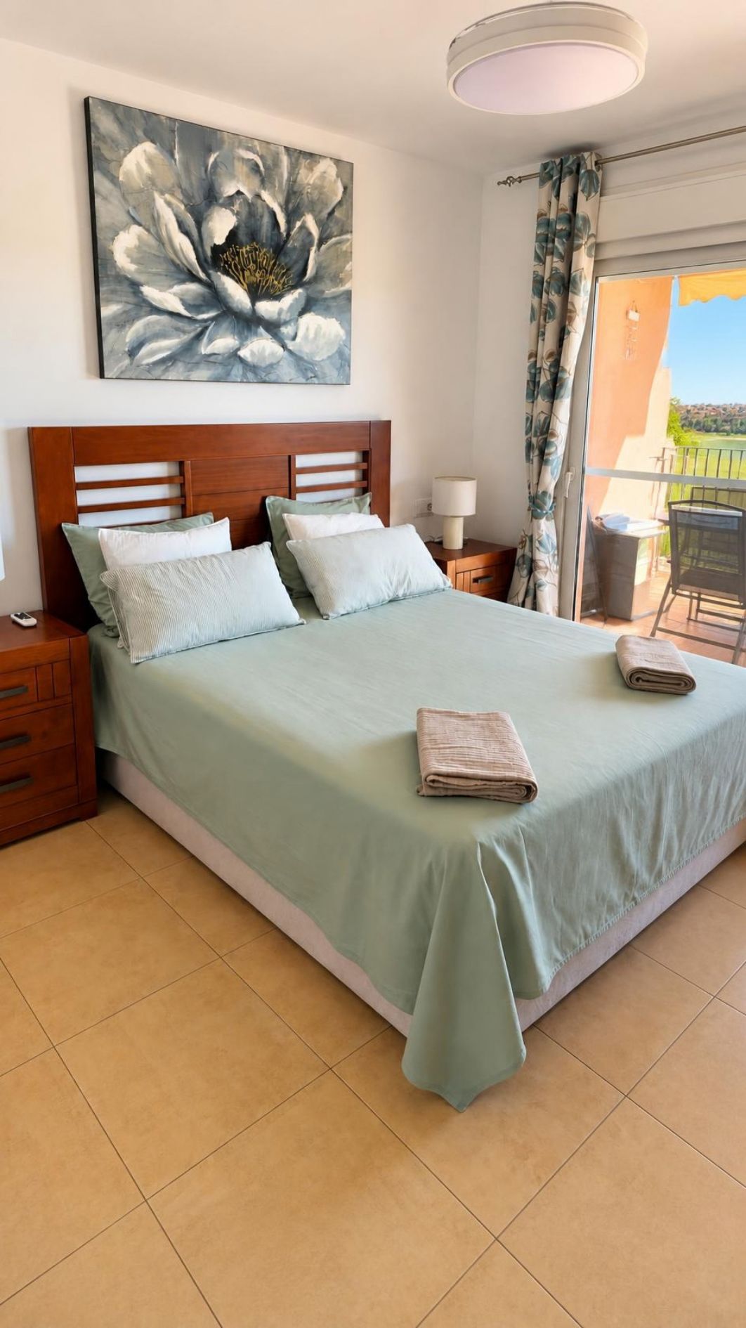 Una cama bien hecha con cubrecama verde claro y almohadas, un cabecero de madera, una mesita de noche con una lámpara y un cuadro de una flor en la pared.