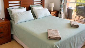 Una cama bien hecha con cubrecama verde claro y almohadas, un cabecero de madera, una mesita de noche con una lámpara y un cuadro de una flor en la pared.
