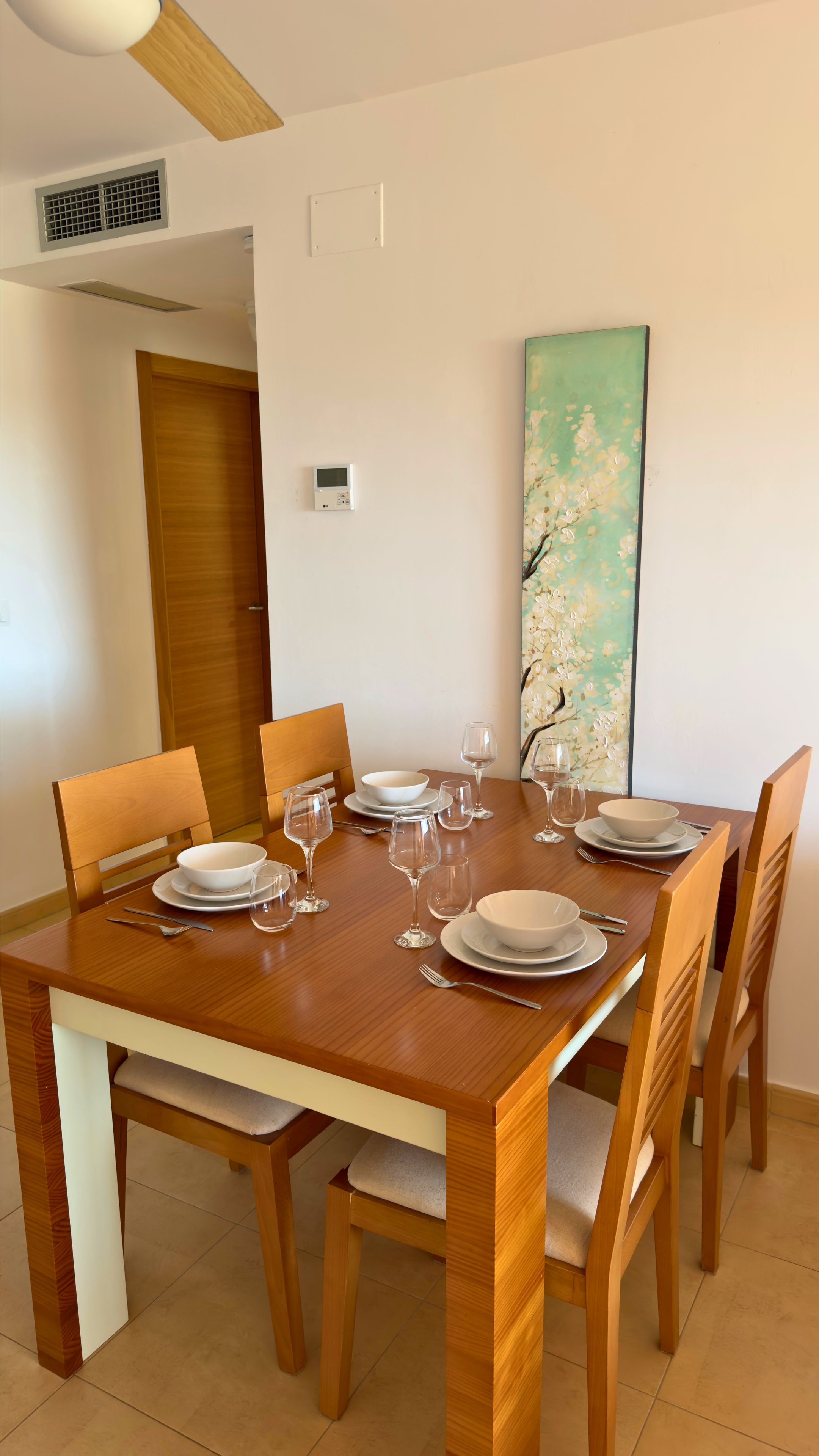 Una mesa de comedor dispuesta con platos, tazones y copas de vino, rodeada de sillas de madera.