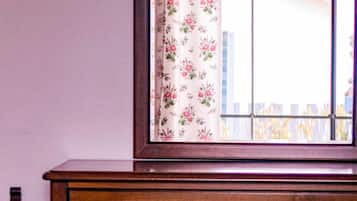 Ein hölzerner Schminktisch mit Spiegel, ein Fenster mit blumigen Vorhängen und ein Bett mit einer blumigen Tagesdecke.