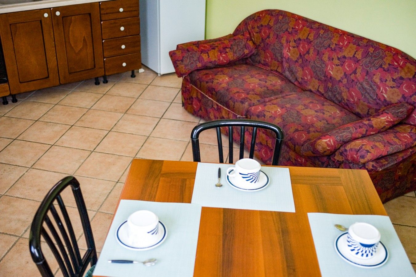 Un comedor con mesa de madera dispuesta para dos personas, dos sillas, un sofá y una zona de cocina con armarios de madera.
