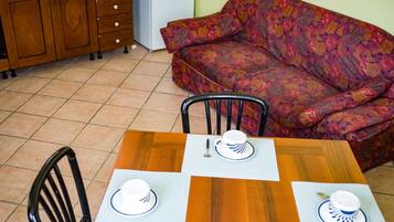 Un comedor con mesa de madera dispuesta para dos personas, dos sillas, un sofá y una zona de cocina con armarios de madera.