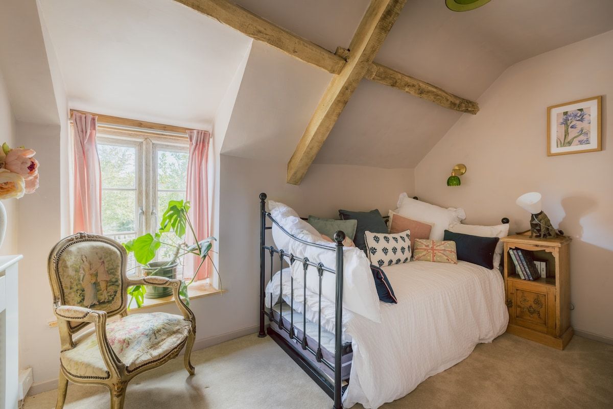 A bedroom with a sloped ceiling, a bed with white bedding, a chair with floral upholstery, and a bedside table with books and a lamp.