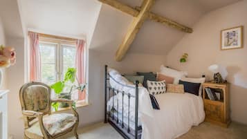 A bedroom with a sloped ceiling, a bed with white bedding, a chair with floral upholstery, and a bedside table with books and a lamp.