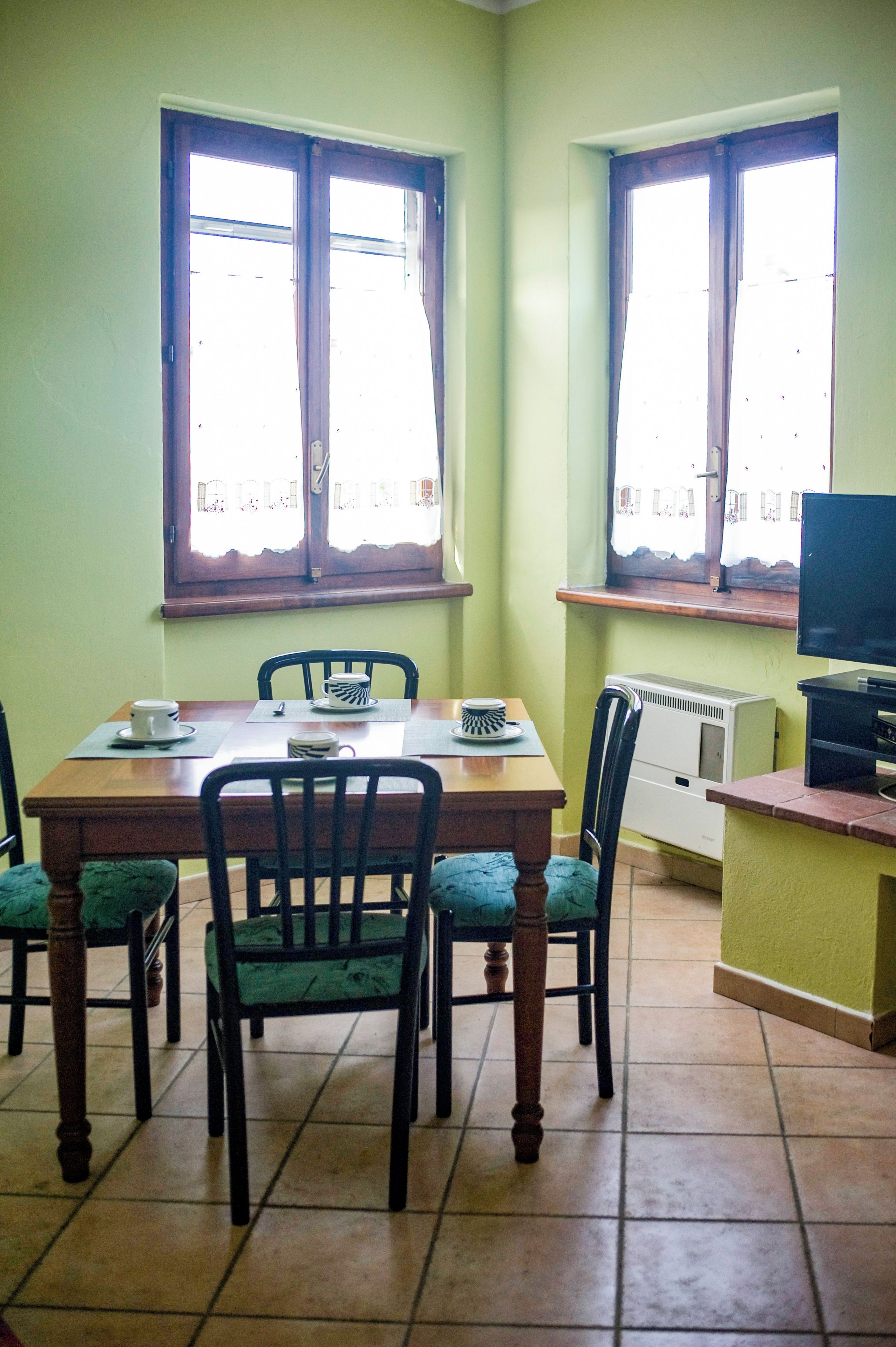 A dining area with a wooden table and chairs, two windows with curtains, and a radiator on the wall.