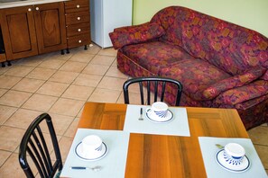 Un coin repas avec une table en bois dressée pour deux personnes, un canapé à motifs floraux et une cuisine en arrière-plan.