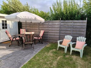 Een terras buiten met een houten tafel en stoelen, een witte parasol en een houten schutting.