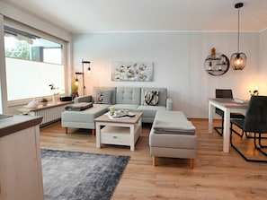 Un salon moderne avec un canapé gris, une table basse, une table à manger avec des chaises et une grande fenêtre laissant entrer la lumière naturelle.