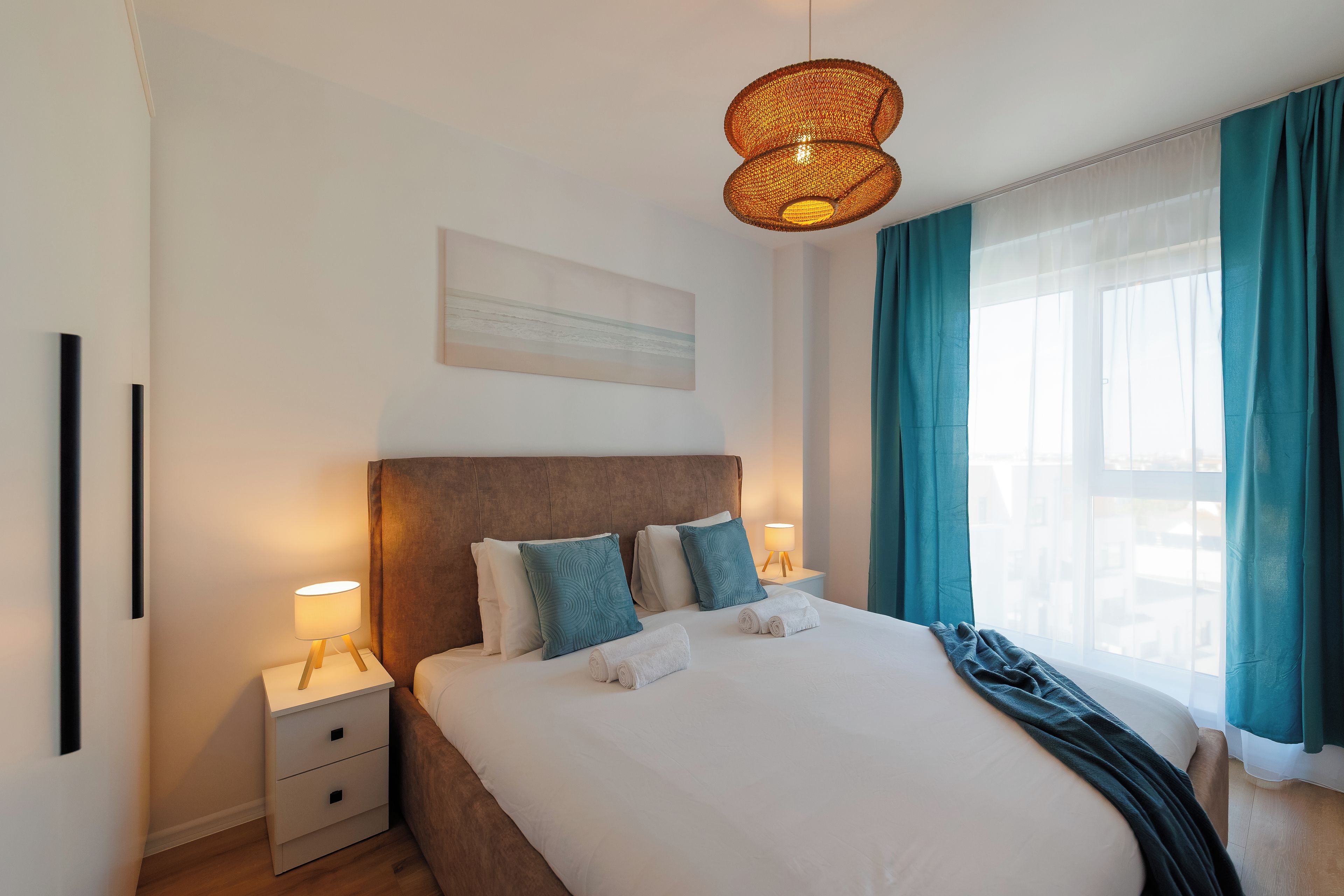 Ett hotellrum med en säng, sänglampor, en sänggavel, ett fönster med gardiner och ett konstverk som hänger på väggen.