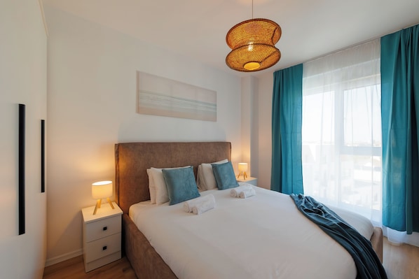 Ett hotellrum med en säng, sänglampor, en sänggavel, ett fönster med gardiner och ett konstverk som hänger på väggen.