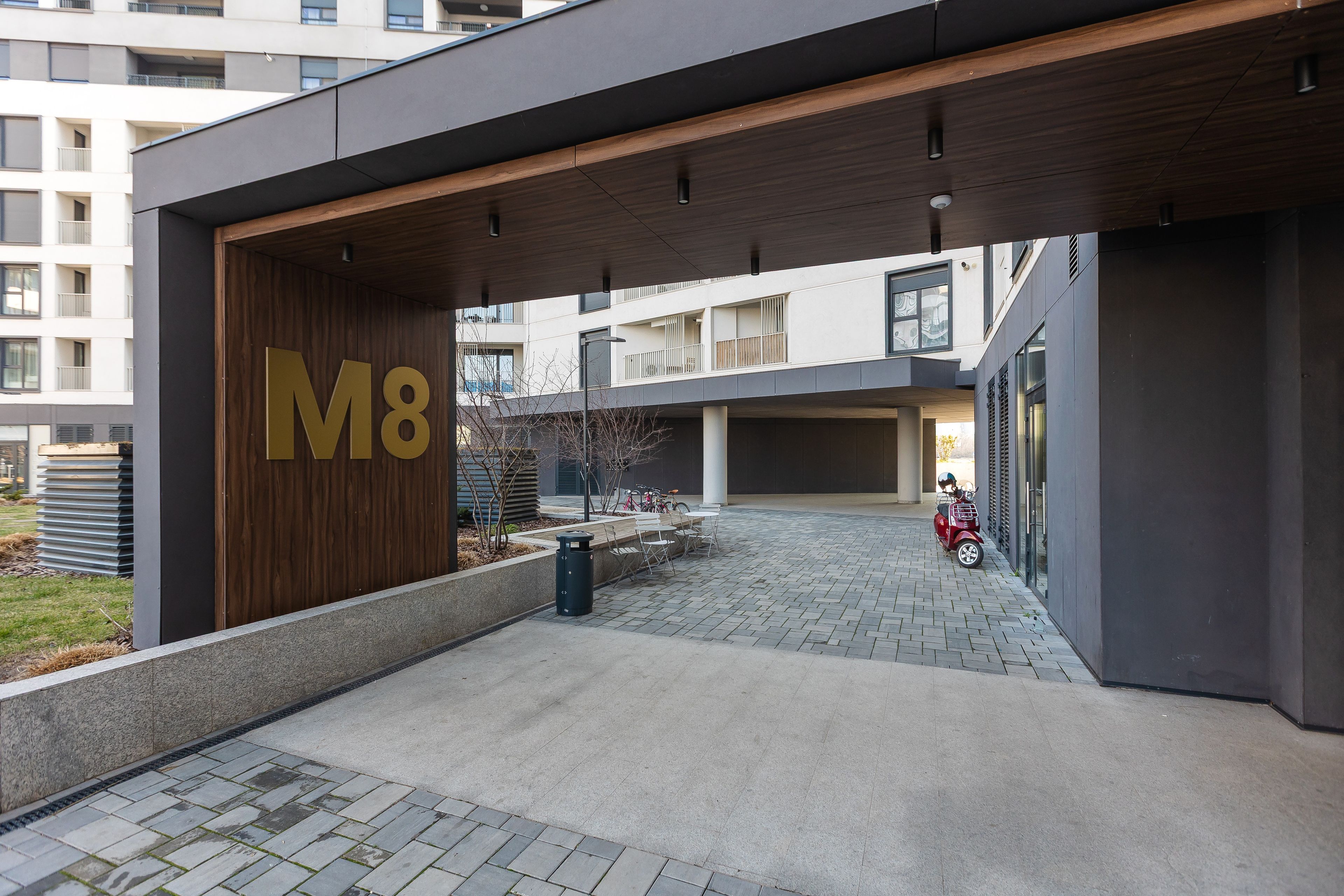 Une entrée moderne avec un grand panneau indiquant « M8 », un espace extérieur couvert avec des sièges et une cour dallée.