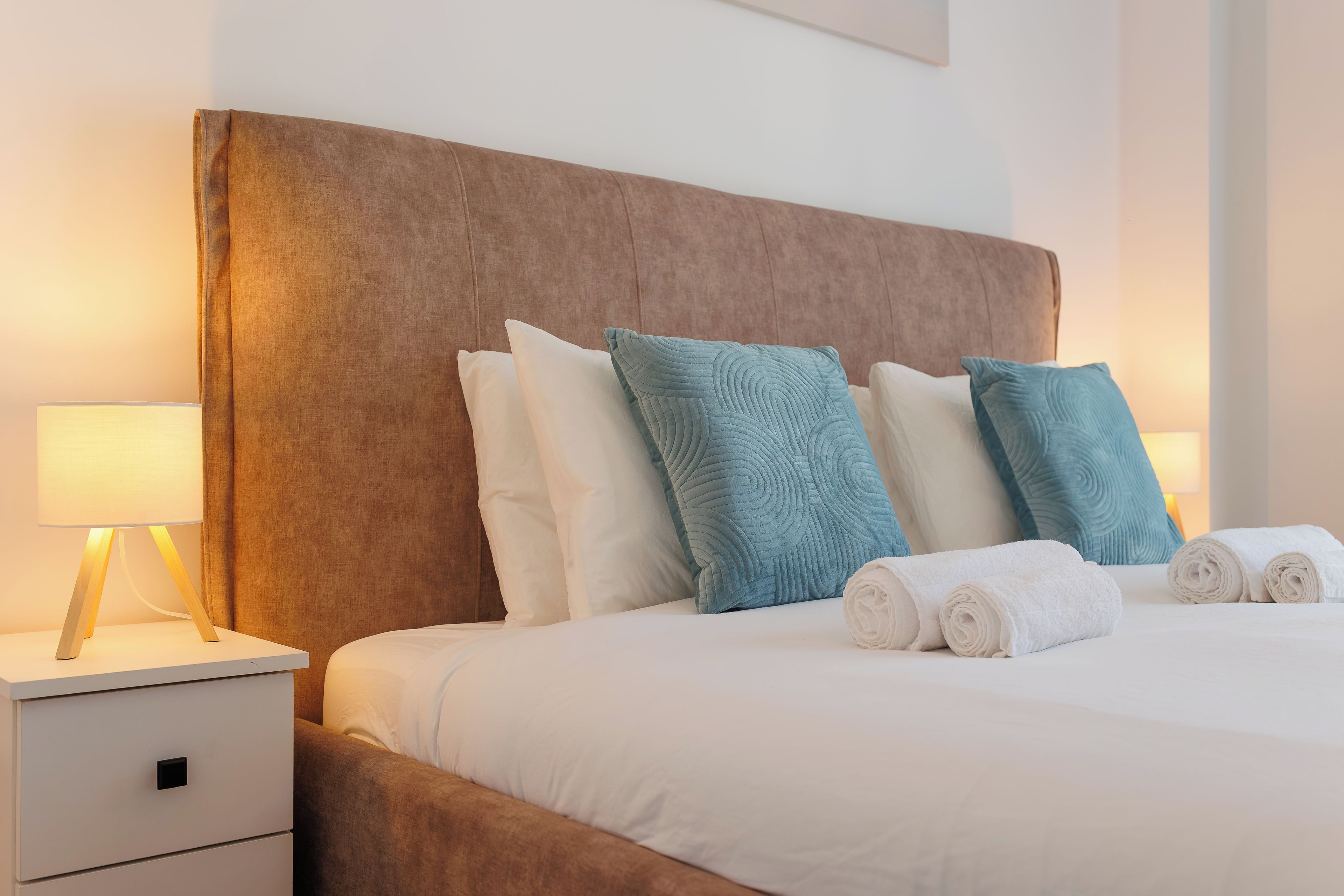 Ein ordentlich bezogenes Bett mit weißen Leinen und zwei Kissen, ein Nachttisch mit einer Lampe und ein Holzbeistelltisch.