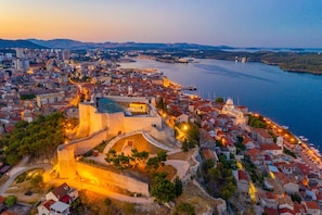 A coastal cityscape with a prominent fortified structure, a harbor with boats, and a mix of residential and commercial buildings. - Villa Nexia With Private Pool (Sibenik)