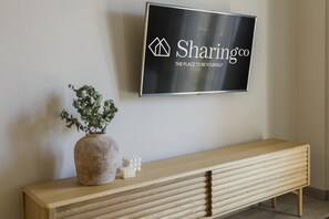 A modern interior with a mounted television displaying "Sharingco" logo, a wooden console table with a vase and decorative item, and a plain wall. - Housing Atarazanas by Sharingco (Málaga)