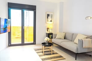Ein modernes Wohnzimmer mit grauem Sofa, einem kleinen Beistelltisch mit Blumenvase, einem gerahmten Bild an der Wand und einer Glastür, die auf einen Balkon führt.