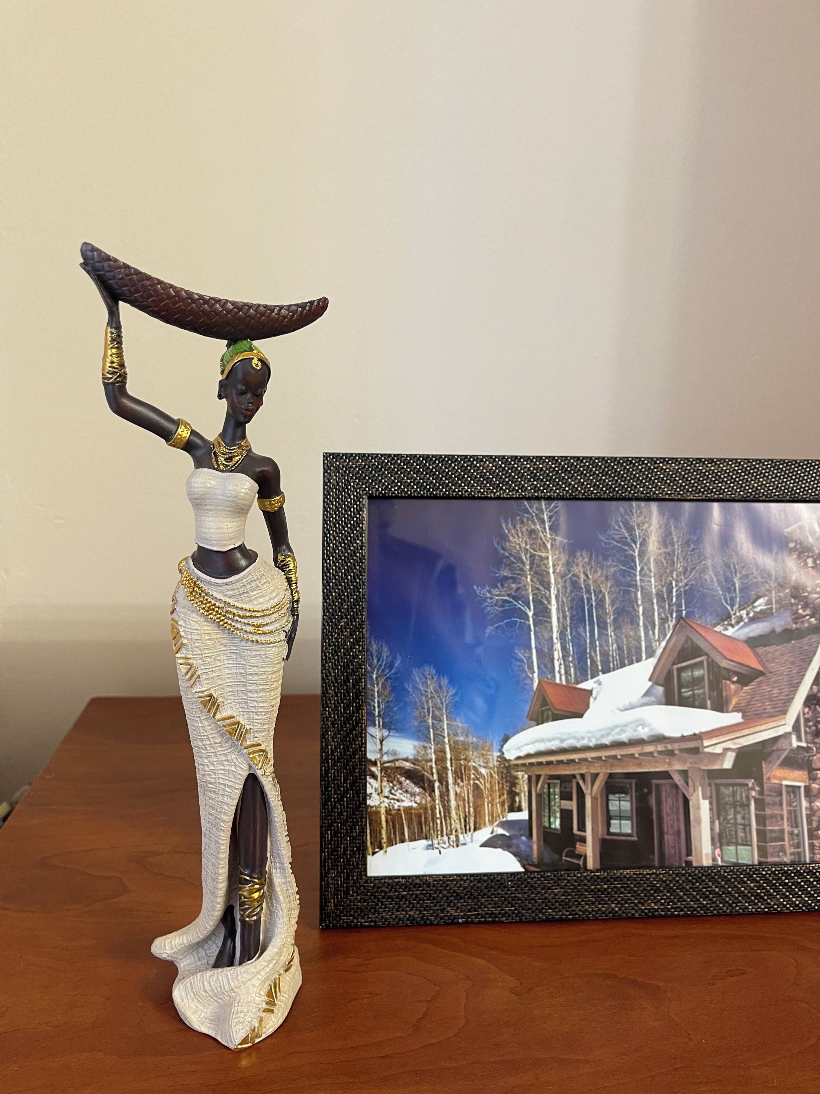 Une sculpture représentant une figure portant un bol sur la tête, à côté d’une photographie encadrée d’une maison se dressant au milieu d’un paysage enneigé.