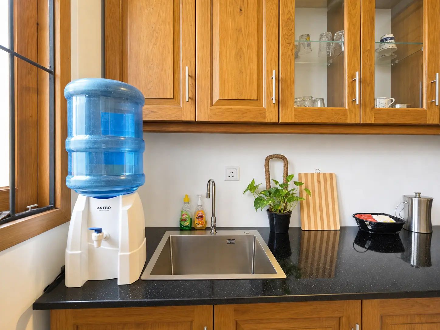 A kitchen with wooden cabinets, a water dispenser, a sink, a potted plant, a cutting board, and a coffee maker.