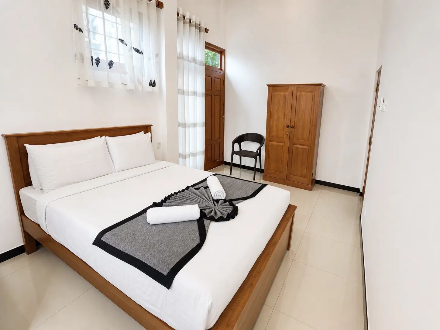 Una cama bien hecha con una manta gris y negra, un armario de madera y una silla en una habitación sencilla con paredes blancas y una ventana con cortinas traslúcidas.