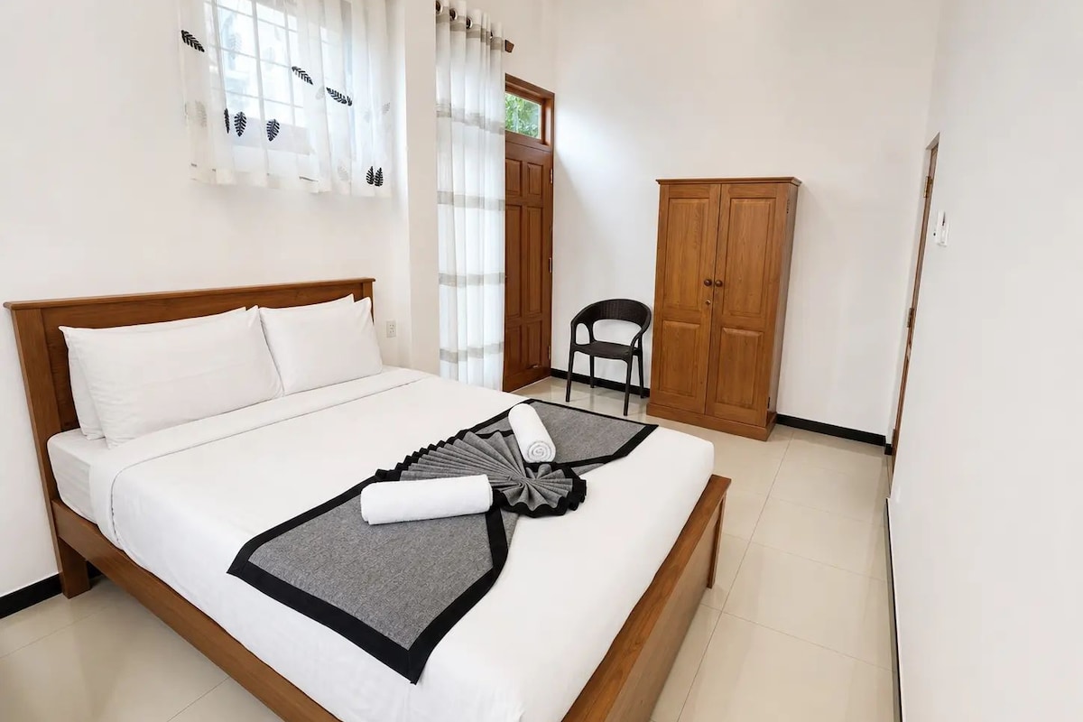 A neatly made bed with a grey and black throw, a wooden wardrobe, and a chair in a simple room with white walls and a window with sheer curtains.