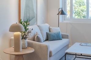 Ein modernes Wohnzimmer mit einem beigefarbenen Sofa, einem runden Holztisch, einer Stehlampe und einem Fenster mit Blick auf Bäume.