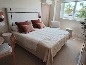 Ein ordentlich eingerichtetes Hotelzimmer mit Bett, Sessel, kleinem Tisch und Fenster mit Blick auf Palmen.