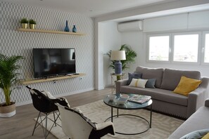 Ein modernes Wohnzimmer mit Flachbildfernseher, einem grauen Sofa mit gelben Kissen, einem Glastisch und einem schwarz-weißen Sessel.