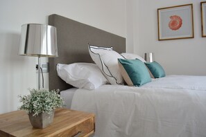 A neatly made bed with white bedding and teal pillows, a bedside lamp, and a small wooden nightstand with a potted plant. - Mlgsa032gra7izq - Calle Granada (Málaga)