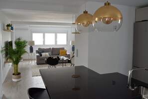 A modern kitchen with a central island, a dining area with chairs, a sofa, and a coffee table. There is a potted plant and a lamp on a side table. - Mlgsa032gra7izq - Calle Granada (Málaga)