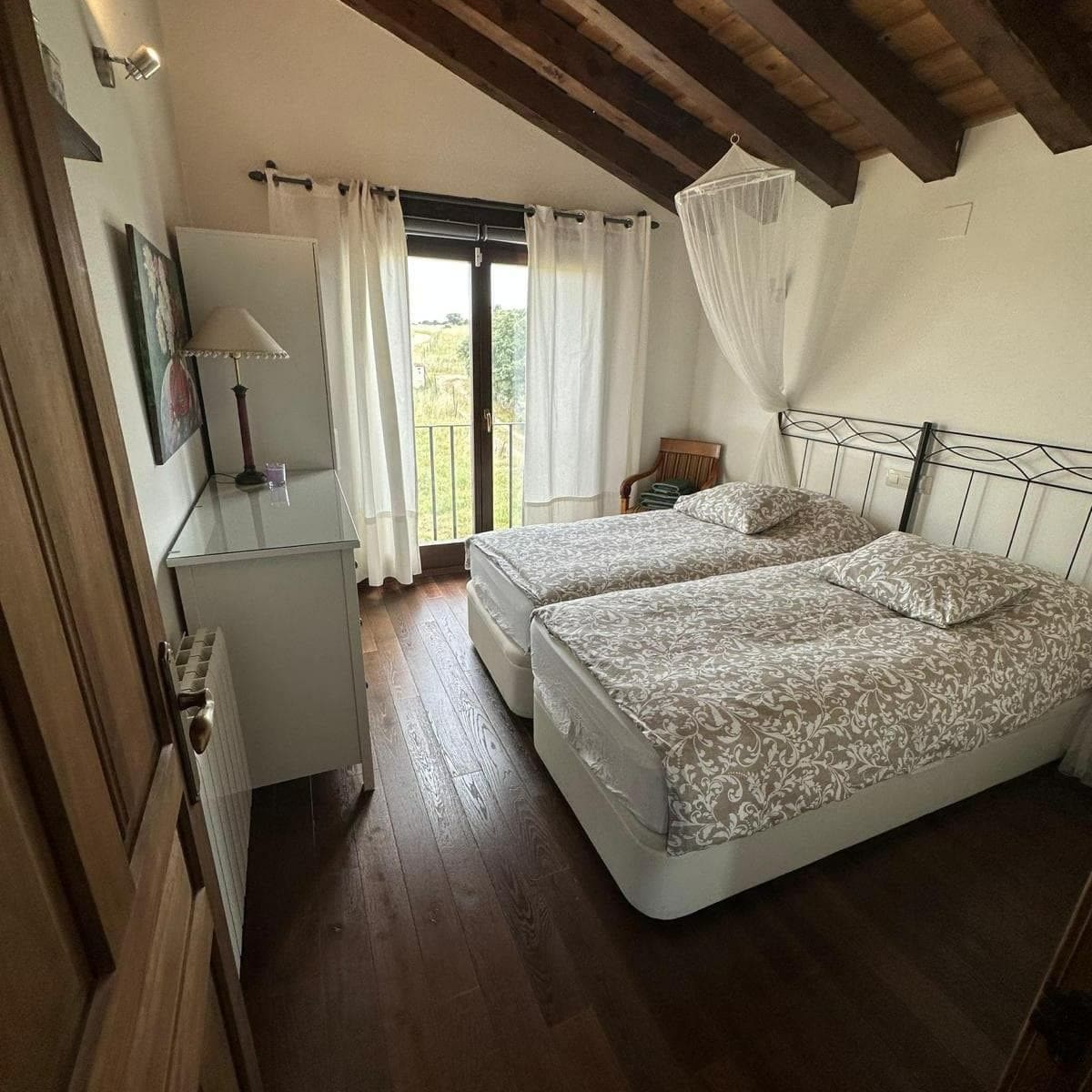 A bedroom with two beds, a small bedside table, a lamp, and a view of the outdoors through a window with curtains.
