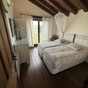 Un dormitorio con dos camas, una mesita de noche pequeña, una lámpara y vistas al exterior a través de una ventana con cortinas.