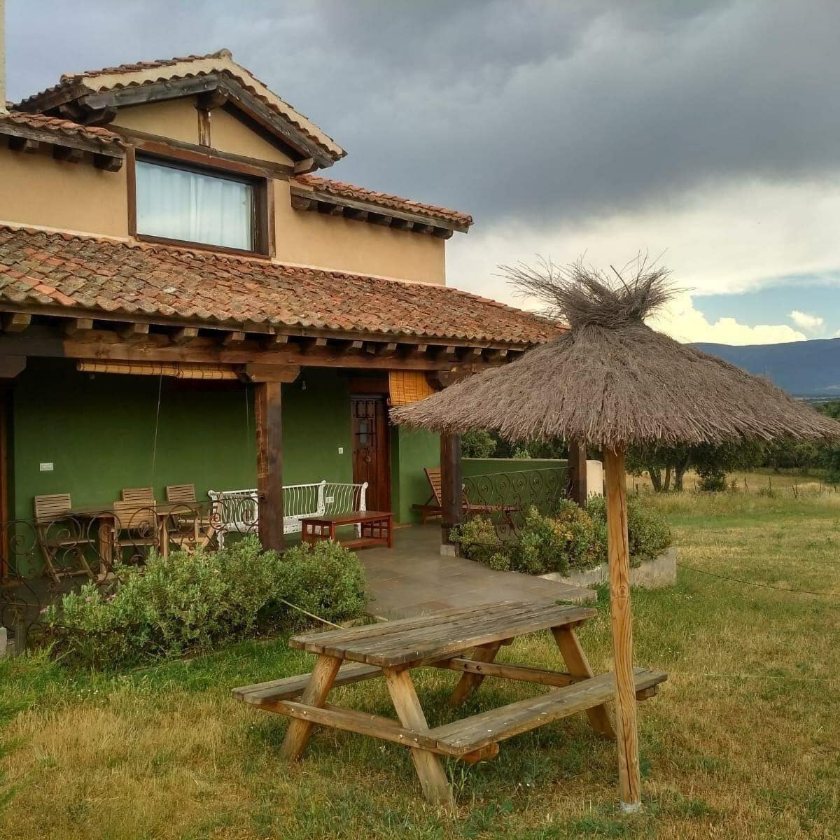 A house with a tiled roof, a thatched roof structure, and a wooden picnic table in the yard.