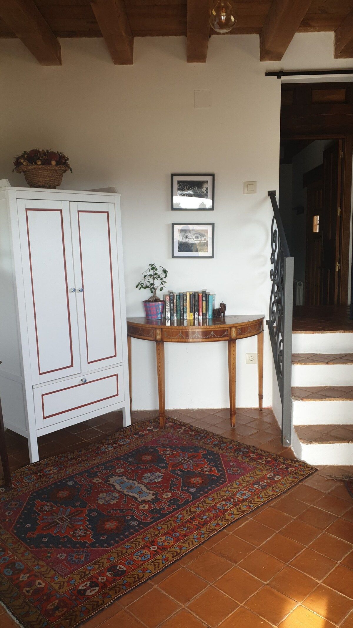 A room with a white cabinet, a wooden table with books and a potted plant, a patterned rug, and a staircase leading to a dark doorway.
