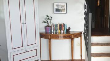 A room with a white cabinet, a wooden table with books and a potted plant, a patterned rug, and a staircase leading to a dark doorway.