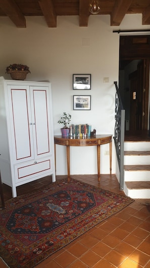 A room with a white cabinet, a wooden table with books and a potted plant, a patterned rug, and a staircase leading to a dark doorway. - Segsa000ntimtt - Segovia (Turegano)