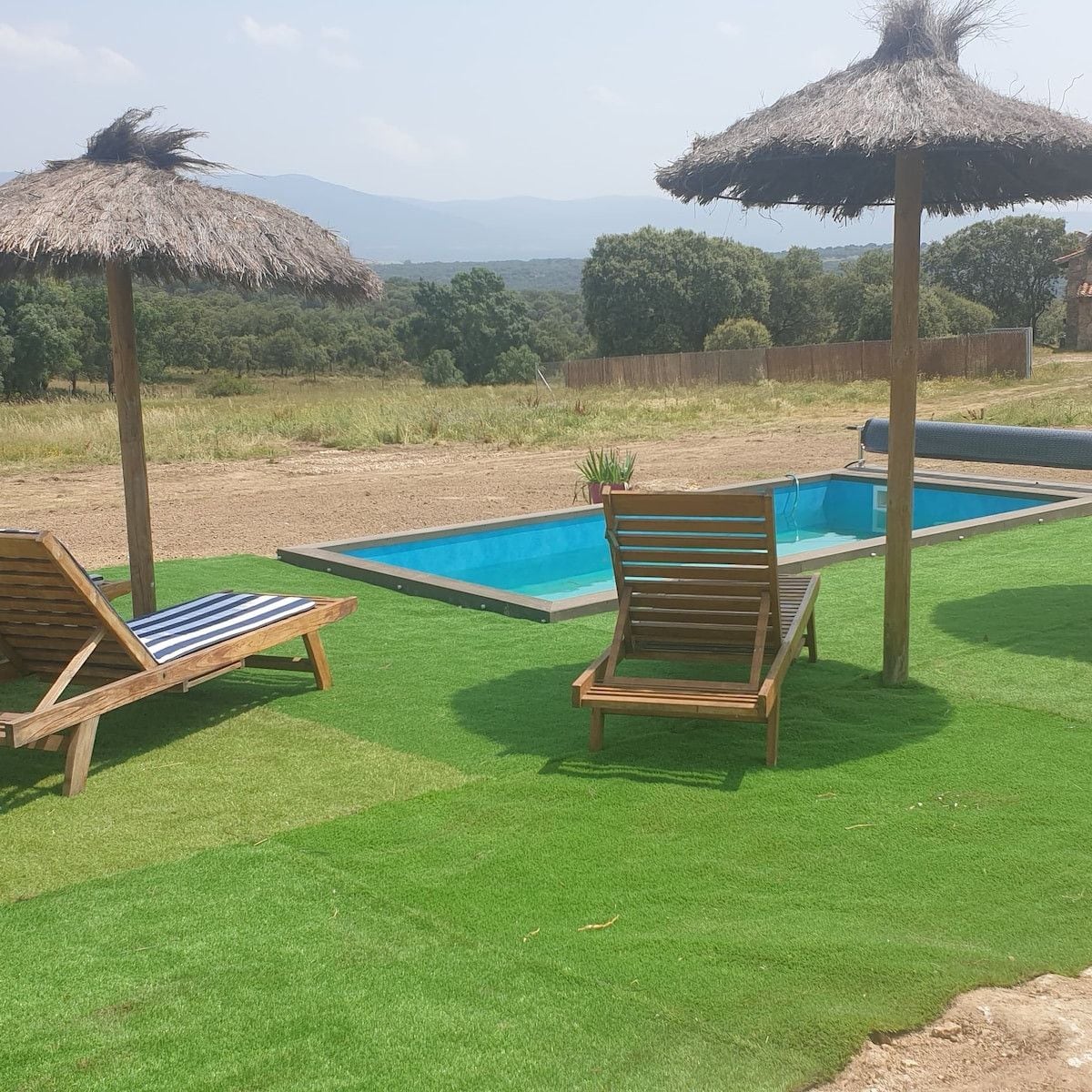 A pool area with two wooden lounge chairs and thatched umbrellas, surrounded by green artificial grass and a view of distant trees and mountains.