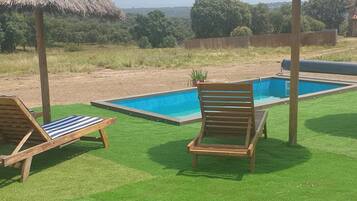 A pool area with two wooden lounge chairs and thatched umbrellas, surrounded by green artificial grass and a view of distant trees and mountains.