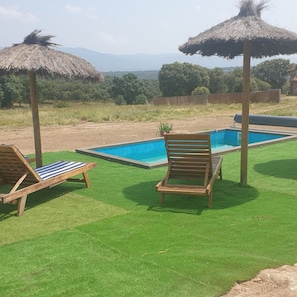 A pool area with two wooden lounge chairs and thatched umbrellas, surrounded by green artificial grass and a view of distant trees and mountains. - Segsa000ntimtt - Segovia (Turegano)