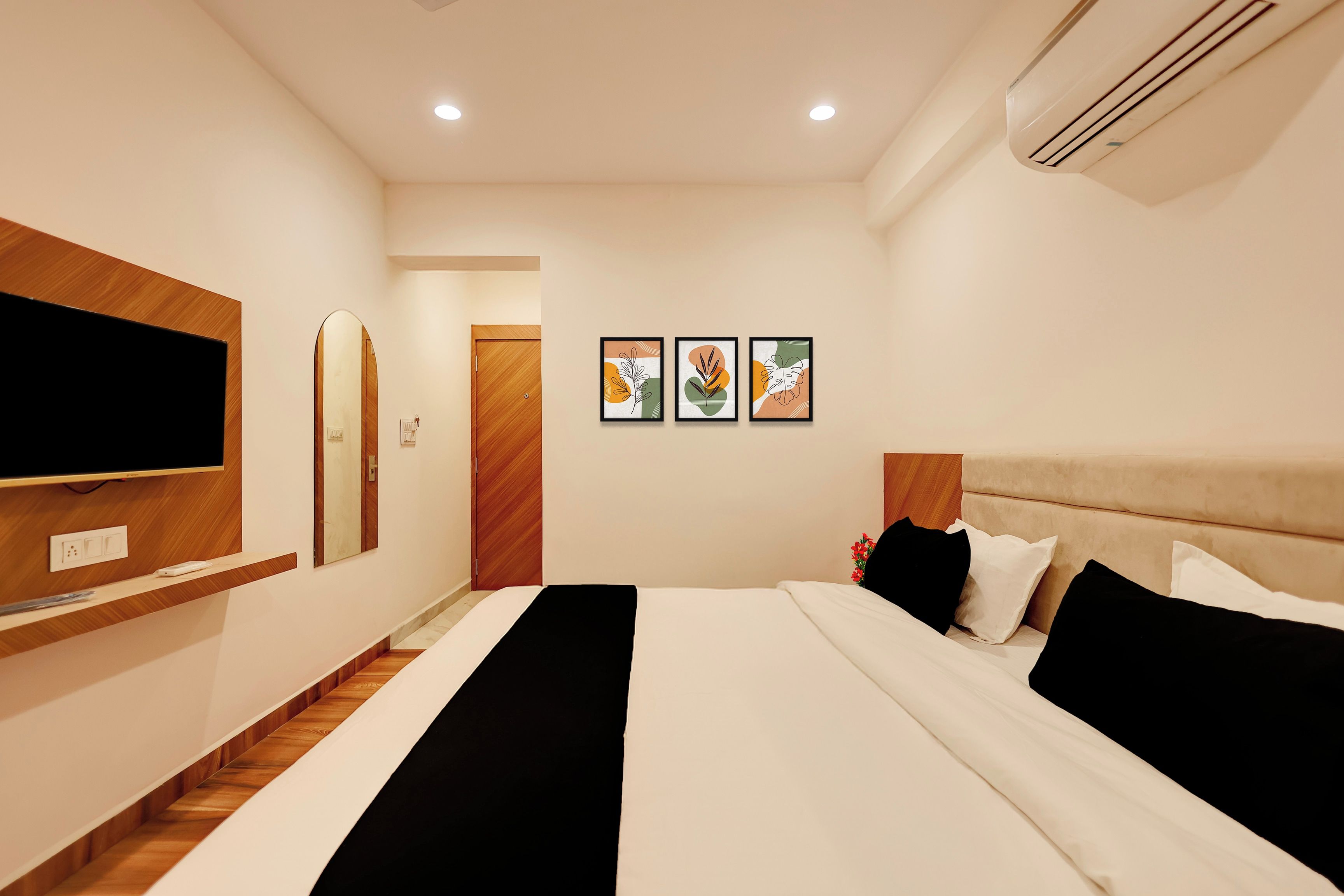 A hotel room with a bed, a flat-screen TV, a mirror, and two framed artworks on the wall.