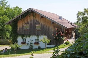 A wooden building with a sloped roof, a porch, and a small garden area with flowers and a tree. - "Kräuterwiese" House, 535 square meters (Frasdorf)